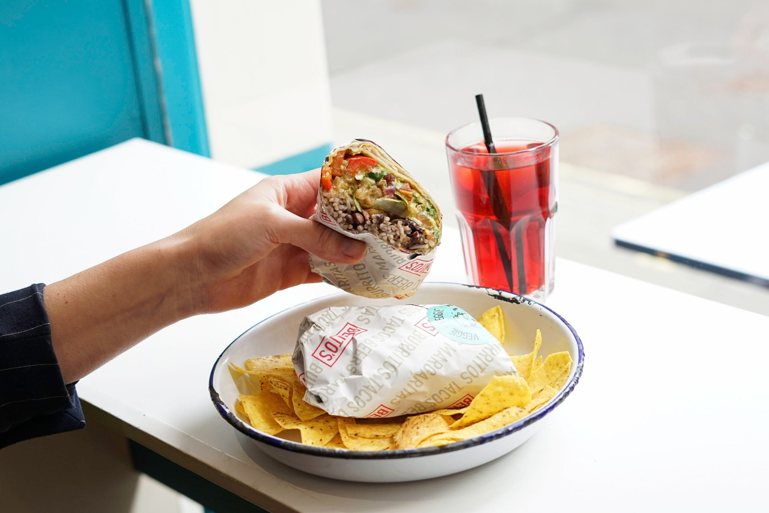 In front of a window, a person holds a burrito and a drink, ready to enjoy their meal.