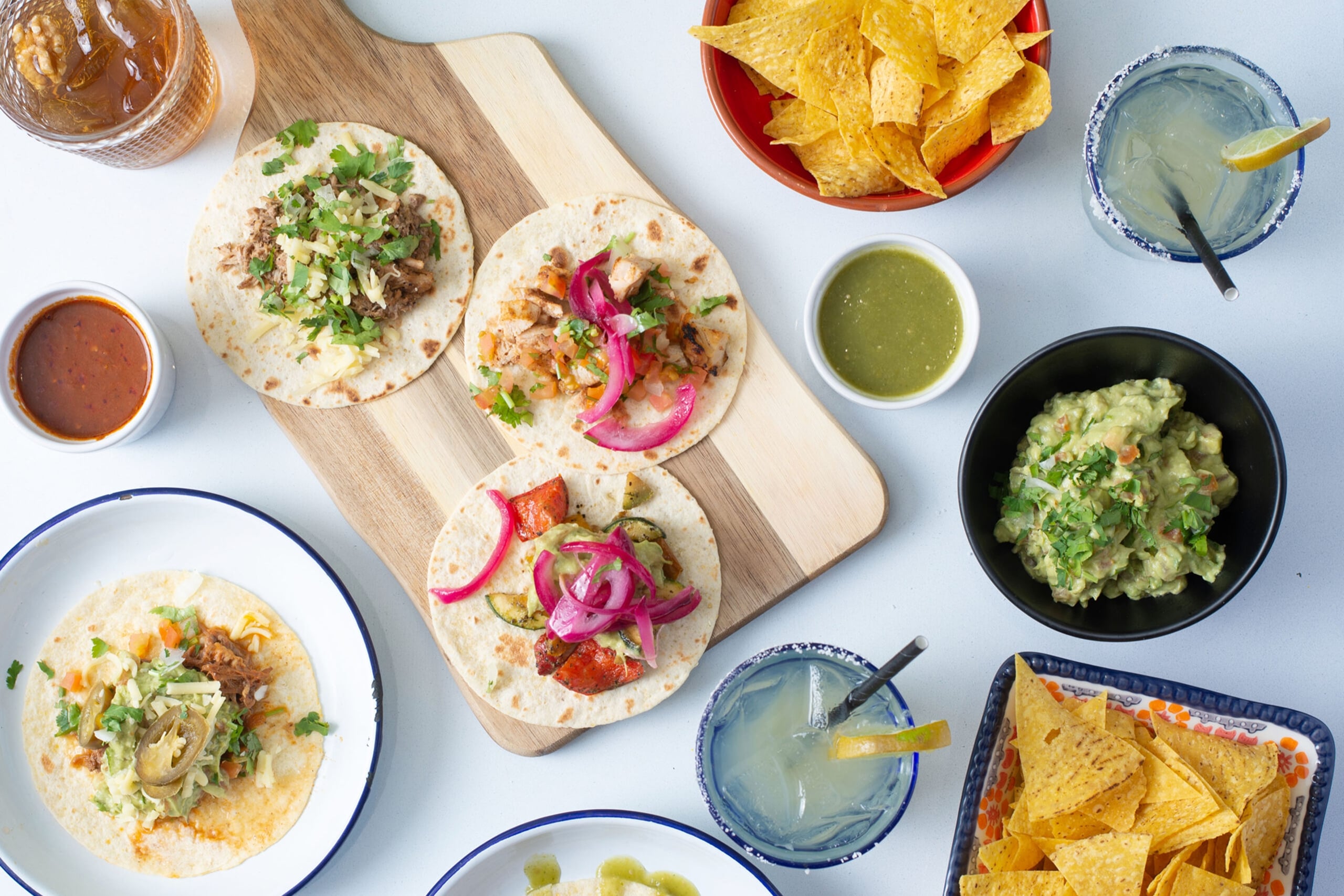 Overhead view of a Mexican meal spread featuring multiple soft tacos on a wooden cutting board with various fillings including meat, pickled red onions, and fresh cilantro. The table includes bowls of guacamole, salsa verde, and red salsa, plates of tortilla chips, two margaritas with lime garnishes, and an additional taco on a white plate with blue rim. All items are arranged on a white surface.