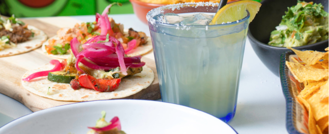 Close-up of a colorful Mexican meal spread featuring soft tacos topped with vibrant ingredients like grilled vegetables and pickled onions, alongside bowls of tortilla chips, guacamole, and a salted-rim margarita with a lemon wedge. A playful green mural serves as the background.