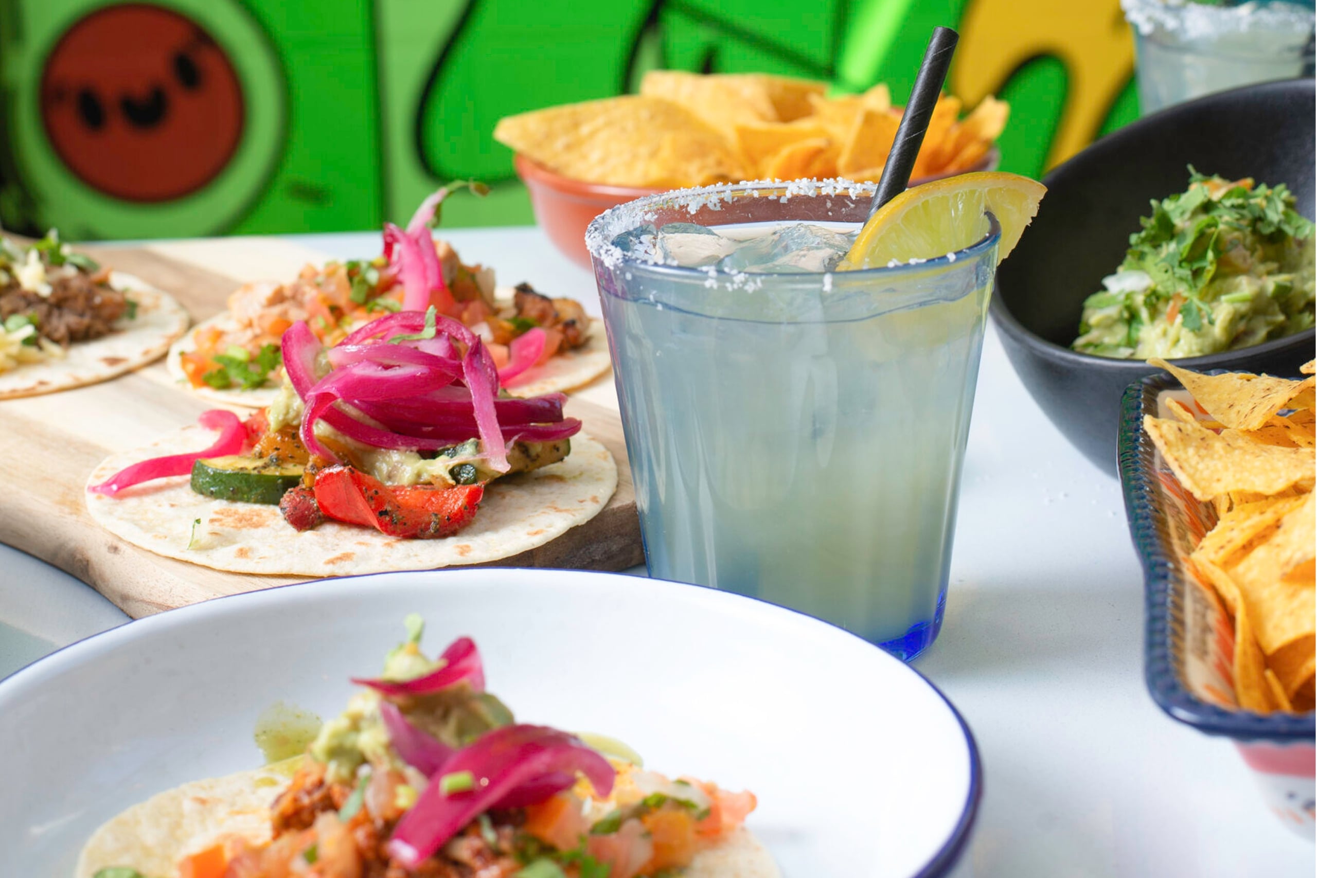 Close-up of a colorful Mexican meal spread featuring soft tacos topped with vibrant ingredients like grilled vegetables and pickled onions, alongside bowls of tortilla chips, guacamole, and a salted-rim margarita with a lemon wedge. A playful green mural serves as the background.